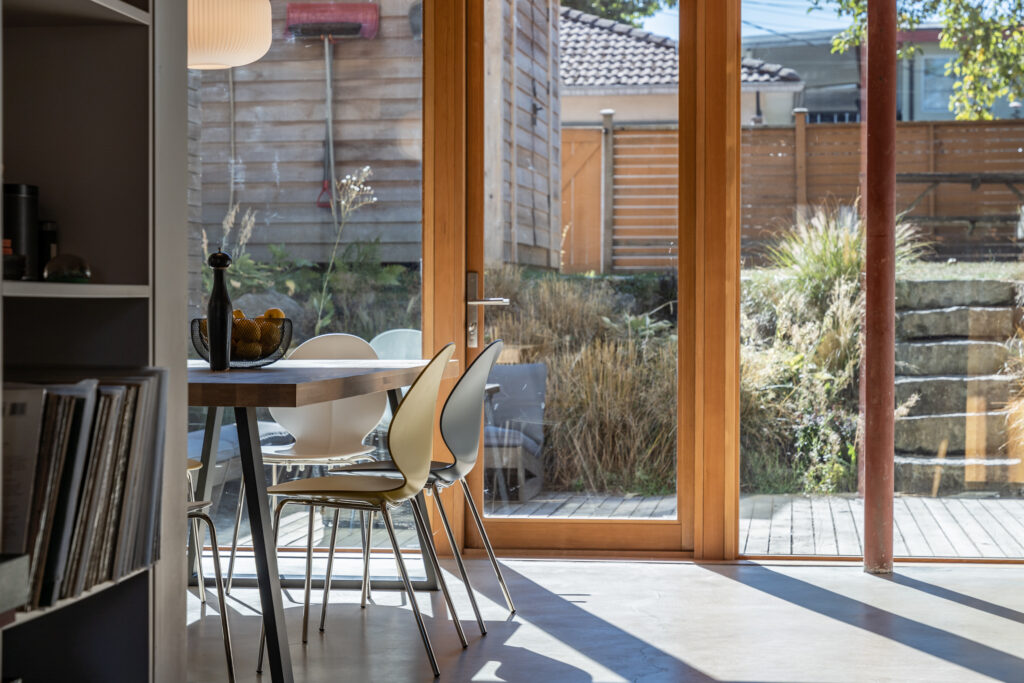 A modern dining area with white chairs and a wooden table is next to large glass doors, overlooking a sunlit garden with stone steps, tall grass, and a wooden fence.