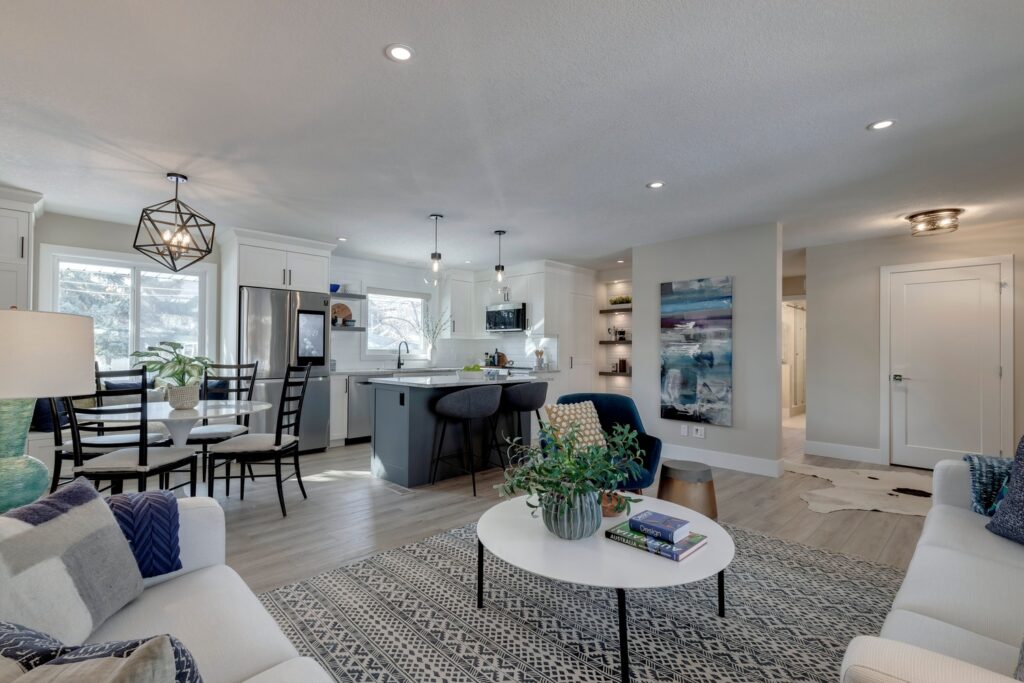 Bright, modern open-concept living area with a white sofa, round coffee table, dining table with black chairs, and a kitchen featuring a center island and pendant lights; large windows let in natural light.