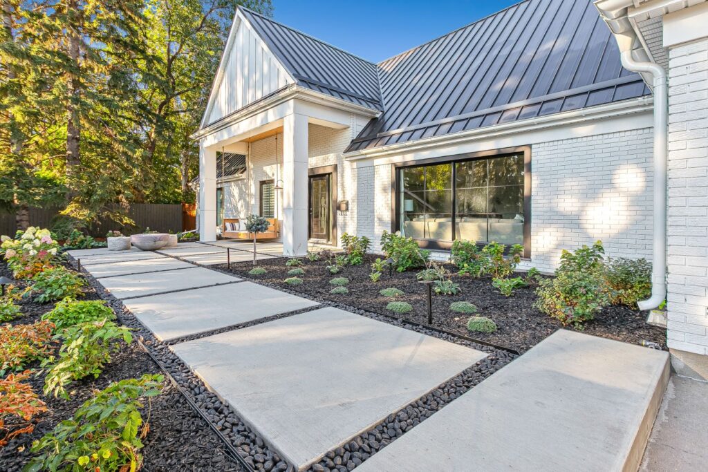 A modern white house with a metal roof, large windows, and a wide concrete walkway bordered by black rocks and neatly landscaped flower beds with small plants and shrubs. Tall trees are visible in the background.