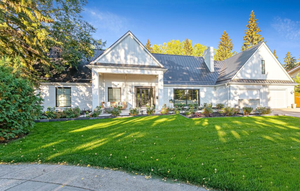 A modern white house with a metal roof, large windows, and double front doors, surrounded by a well-maintained lawn, colorful landscaping, and tall trees under a clear blue sky.