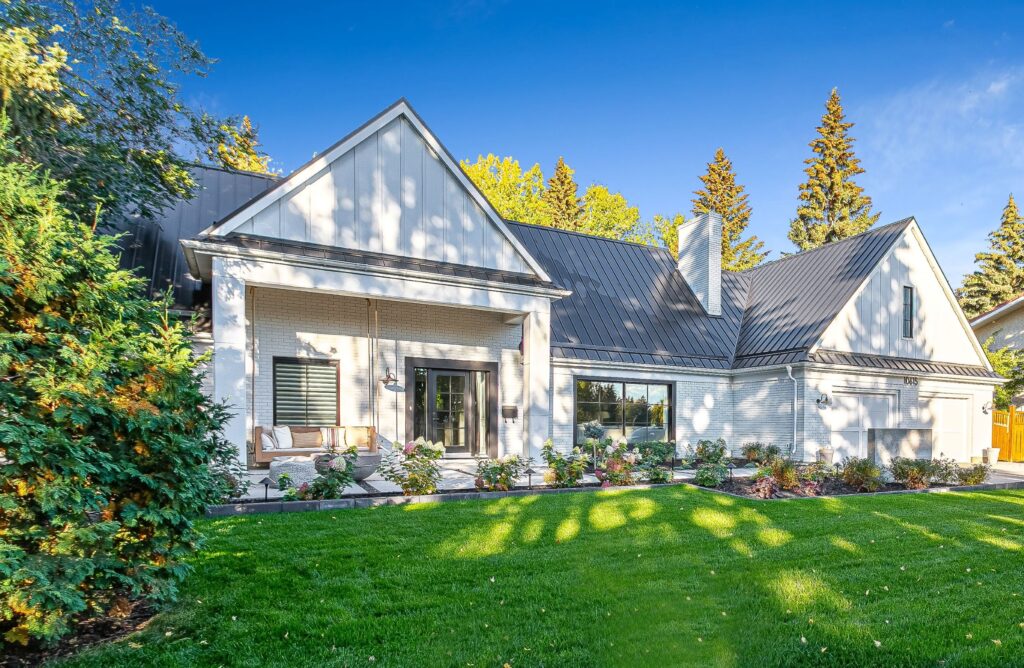 A modern white house with a dark metal roof, large windows, and a covered porch. The home is surrounded by green grass, landscaped shrubs, and tall trees under a clear blue sky.