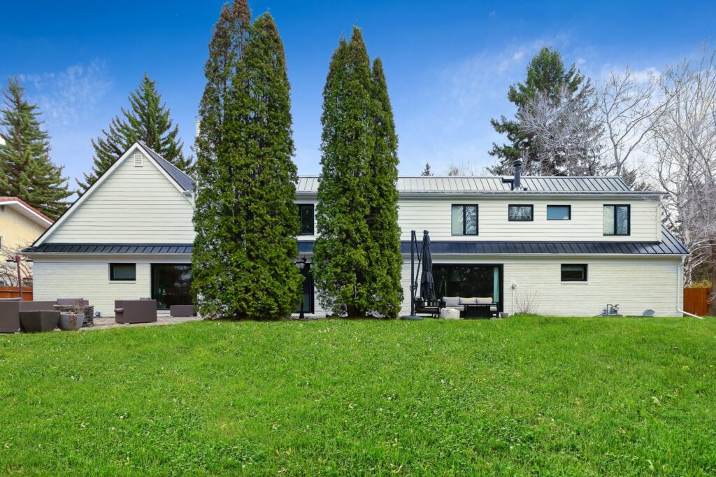 A modern, white two-story house with large windows and a dark metal roof, set behind two tall evergreen trees, with a spacious green lawn in the foreground and a clear blue sky above.