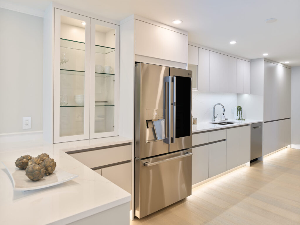 Modern kitchen with light-colored cabinets, stainless steel refrigerator, glass-front display cabinet, built-in sink, under-cabinet lighting, and white countertops; decorative artichokes on a plate in the foreground.