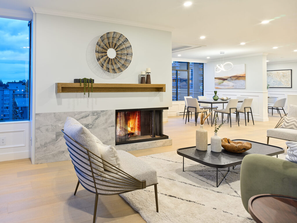 A modern, open-concept living and dining area with light wood floors, a marble fireplace, contemporary furniture, and large windows showing a city view at dusk. Neutral tones and minimalist decor create a cozy, elegant atmosphere.