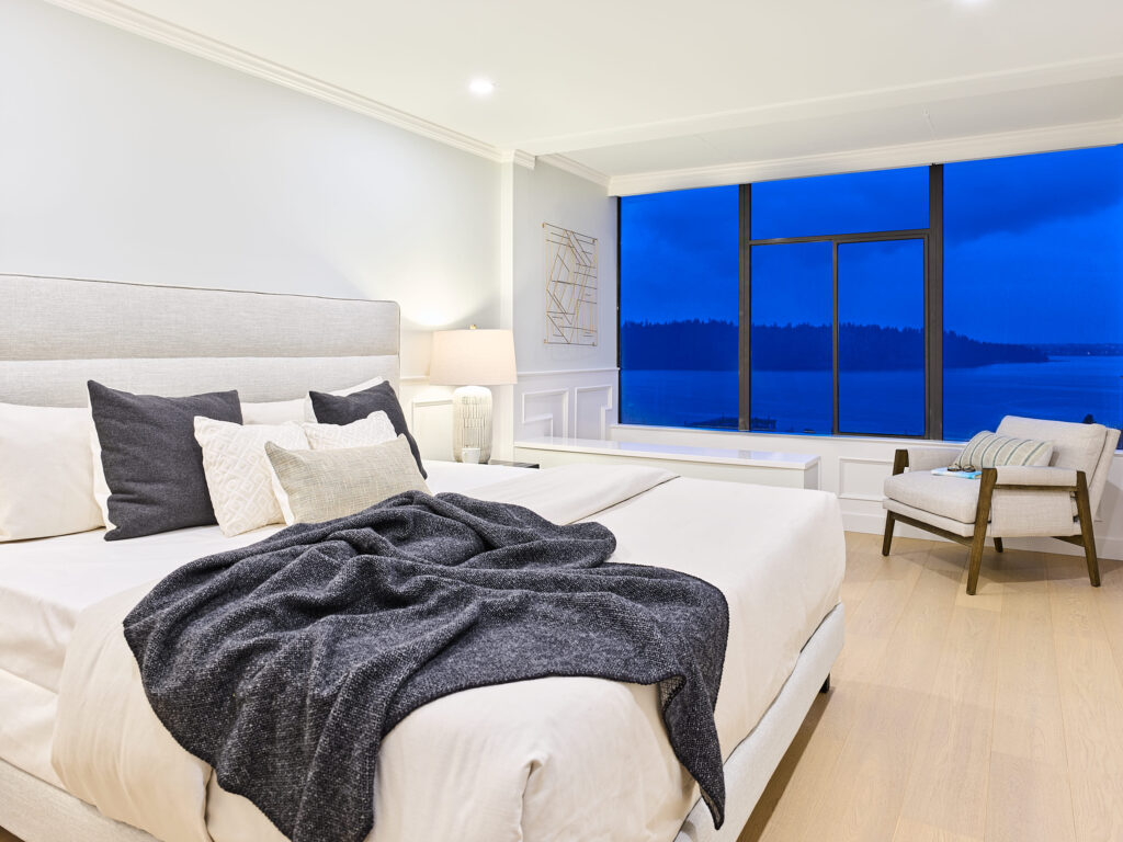 Modern bedroom with a large, neatly made bed, cozy pillows, and a dark throw blanket. A lounge chair sits by a floor-to-ceiling window with a view of a lake and trees at dusk. The room is softly lit and spacious.