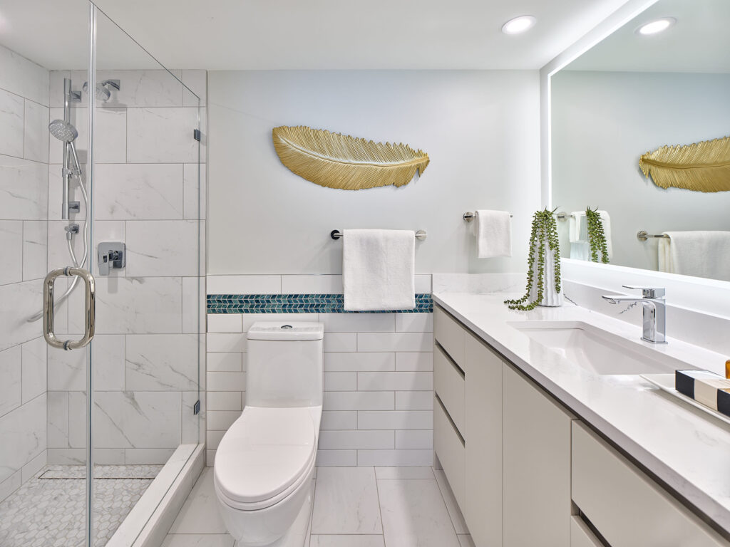 Modern bathroom with a glass-enclosed shower, white tiles, a toilet, a long white vanity with a sink, a potted plant, and a gold leaf wall decoration above a towel rack.