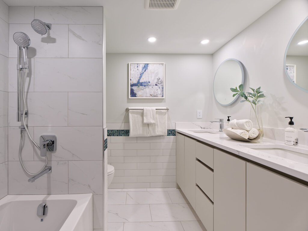 Modern bathroom with a white tub and shower, large vanity with double sinks, round mirrors, white cabinets, rolled towels, a potted plant, and wall art above a towel rack, all in a neutral color palette.