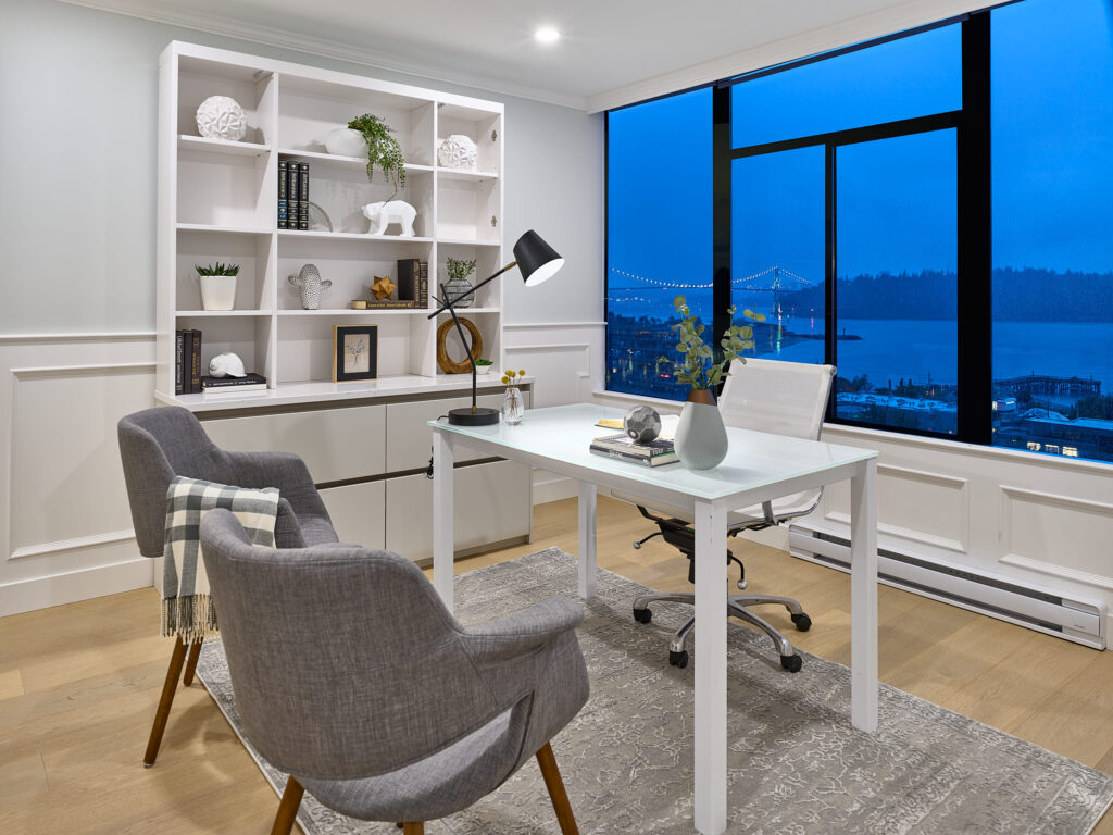 Modern home office with a white desk, two gray chairs, shelves with decor, and a large window overlooking a city skyline and bridge at dusk. A desk lamp, books, and plants add cozy touches.