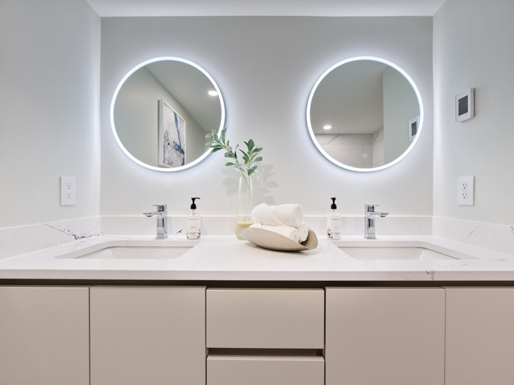 A modern bathroom with two sinks, circular backlit mirrors, a plant in a vase, and rolled towels on a white marble countertop. The space is clean and minimalistic with soft, neutral colors.
