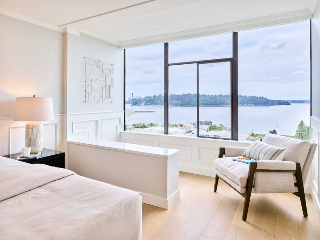 A bright, modern bedroom with a large window offering a scenic view of water and trees. The room features a cozy armchair, a neatly made bed, a nightstand with a lamp, and abstract wall art.