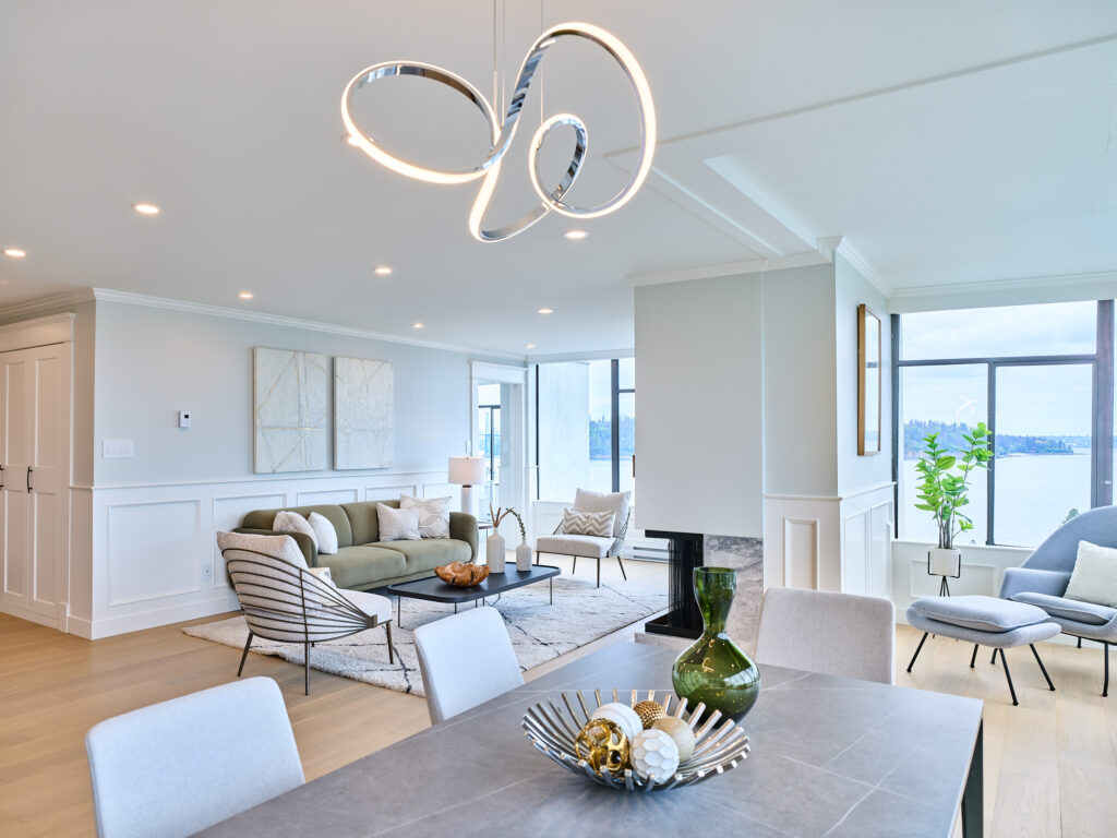 Modern, bright living and dining area with large windows, neutral furnishings, contemporary lighting, and light wood floors. Decorative pieces are on the dining table, and there is a view of water and greenery outside.