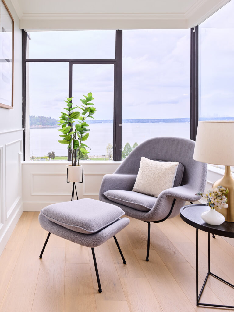 A modern lounge chair with a matching ottoman sits by large windows overlooking a lake. A side table with a lamp and flowers, a potted plant, and light wood floors create a bright and serene reading corner.