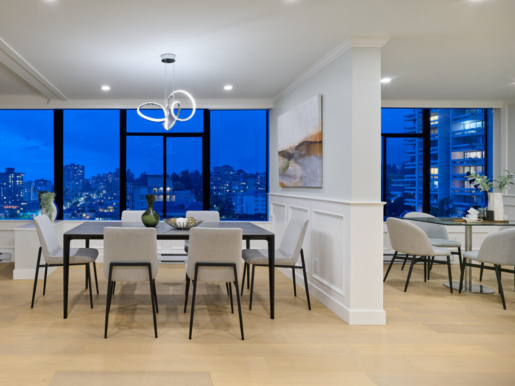 Modern dining room with a black table, eight light gray chairs, contemporary chandelier, city view through large windows, light wood flooring, and abstract wall art; adjacent to a sitting area with a small table.