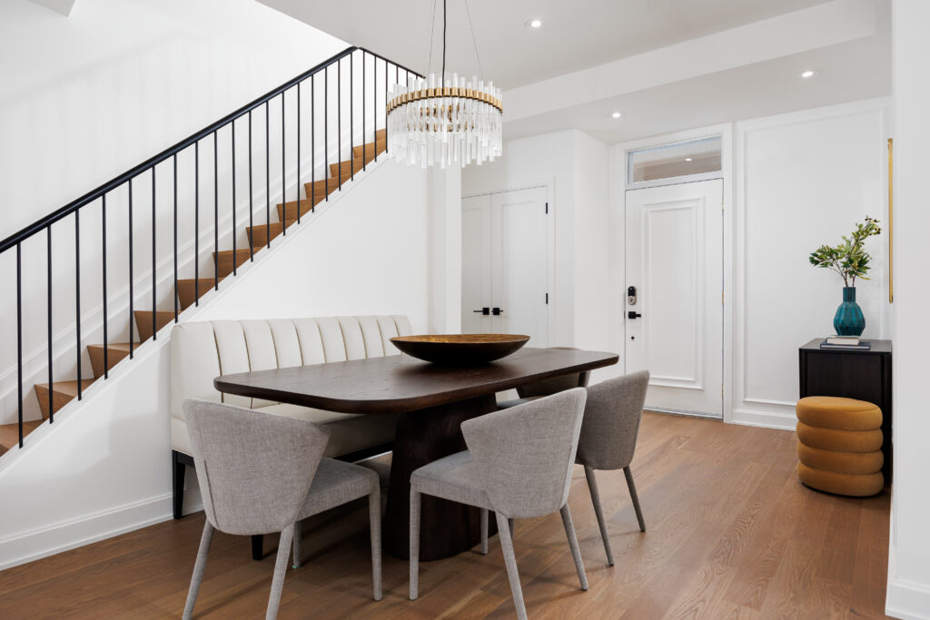Modern dining room with a dark wood table, six light gray chairs, a white upholstered bench, chandelier, wood stairs with black railing, and a small side table with a blue vase and yellow ottoman near the white front door.