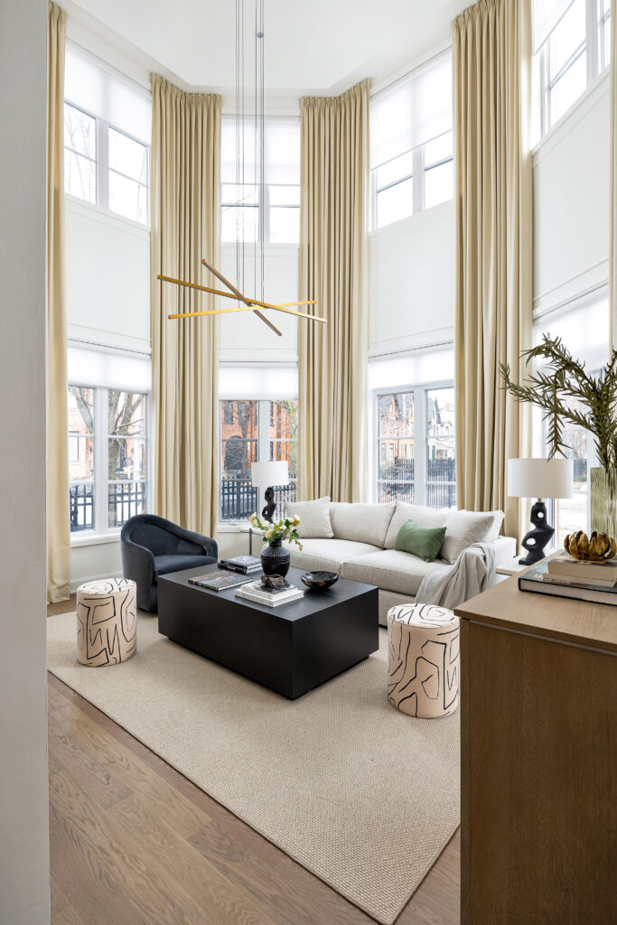 A modern living room with tall windows and beige curtains, a light gray sofa, a black armchair, a rectangular black coffee table, two abstract-patterned stools, and a neutral rug on wood flooring.