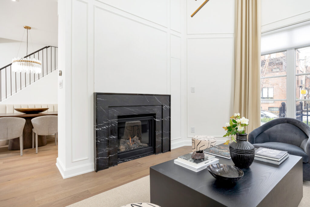 A modern living room with a black marble fireplace, light wood flooring, a black coffee table with decor, beige curtains, and large windows. In the background, a dining area with a round table and upholstered chairs is visible.