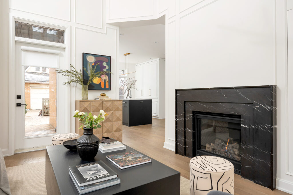 A modern living room with a black marble fireplace, a black coffee table with books and a vase of flowers, patterned stools, abstract artwork, and a view into a bright kitchen and outdoor area.