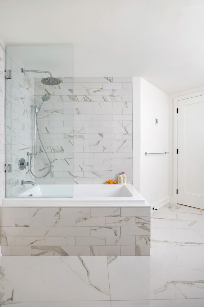A modern bathroom with a white bathtub, marble tile surround, glass shower screen, rainfall showerhead, and a few bath products on the tub edge. The floor and walls are also tiled in white marble.