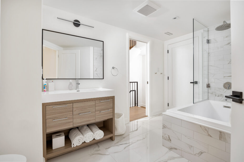 Modern bathroom with white walls, a large mirror above a wooden vanity with double sink, neatly rolled towels, a glass-enclosed shower, and a bathtub with marble tiles. Doorway leads to a bright hallway.