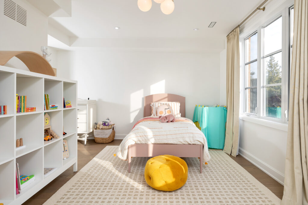 A bright, tidy childs bedroom with a single pink bed, white shelving unit with toys, a yellow pouf, and large windows with beige curtains letting in natural light.