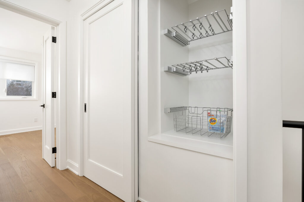 An open closet with wire shelving and a metal basket containing a single box of Tide detergent. The closet is in a bright hallway with wood floors and white walls. Two closed doors and a lit room are visible nearby.