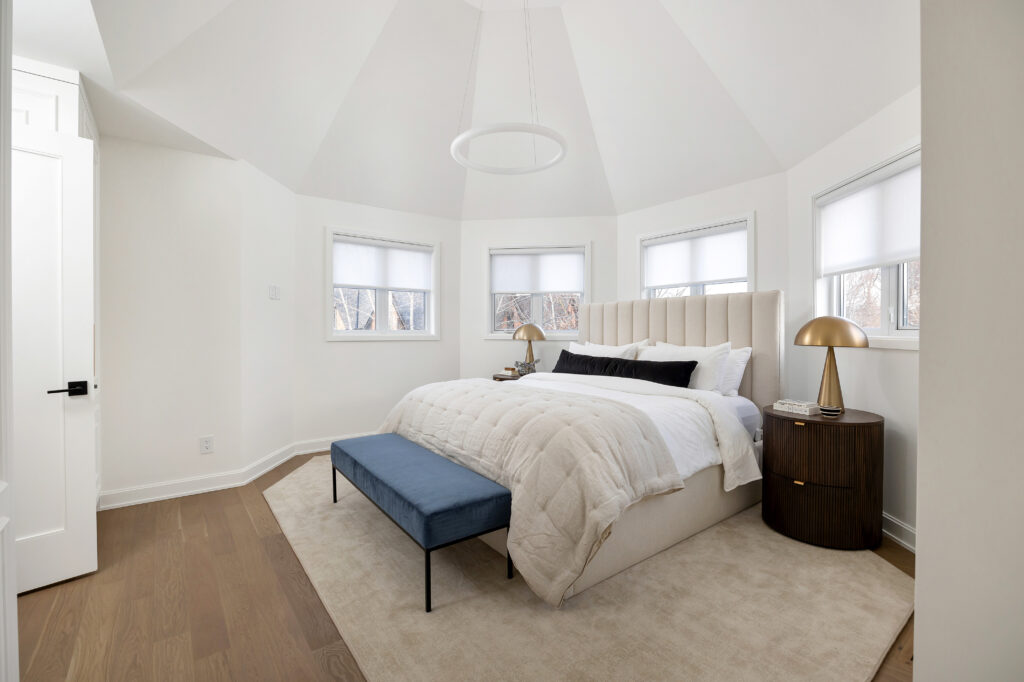 A modern, white bedroom with a tall, tufted headboard, beige bedding, a blue bench at the foot of the bed, gold bedside lamps on round wood tables, hardwood floors, and multiple windows with white shades.