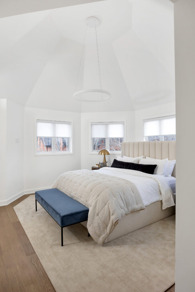 A modern, bright bedroom with white walls, a beige upholstered bed, white bedding, black and white accent pillows, a blue bench at the foot of the bed, and three windows with white shades.