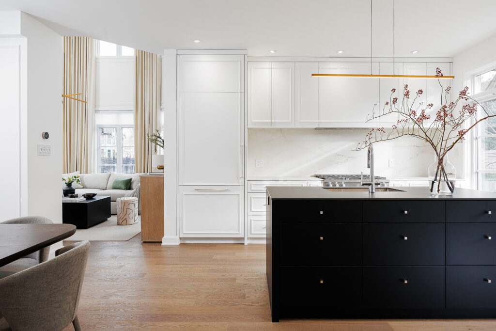 A modern kitchen with white cabinets, a marble backsplash, and a black island. Wooden floors and neutral decor create a bright, open space. A cozy living area with a sofa is visible in the background near large windows.