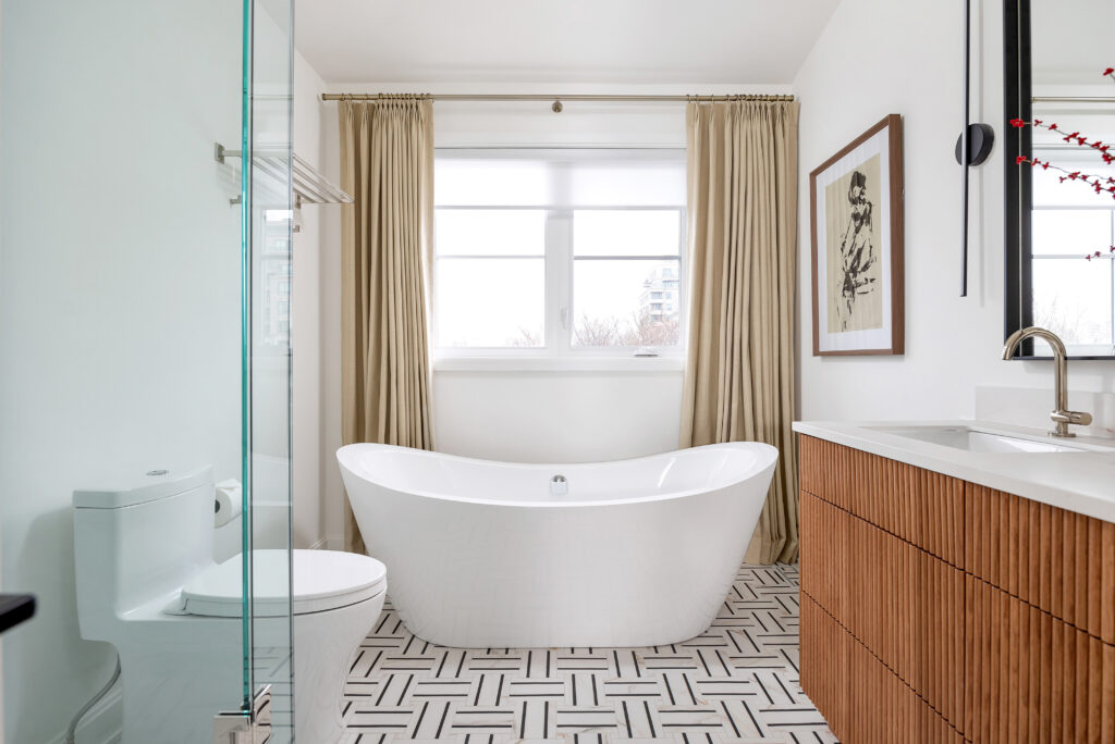 Modern bathroom with a freestanding white bathtub in front of a large window with beige curtains, geometric tile floor, wood vanity, and a wall-mounted faucet next to an artistic framed print.