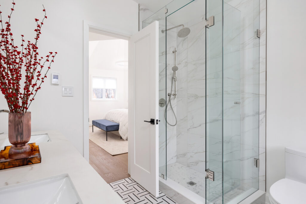 Modern bathroom with a glass shower, marble walls, white countertops, a vase with red branches on the counter, and a view into a bright bedroom with a blue bench and white rug.