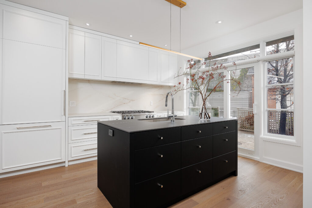 Modern kitchen with white cabinets, light wood flooring, a black island with drawers, a built-in oven, and a vase with branches. Large windows let in natural light and offer a view of a patio and trees outside.