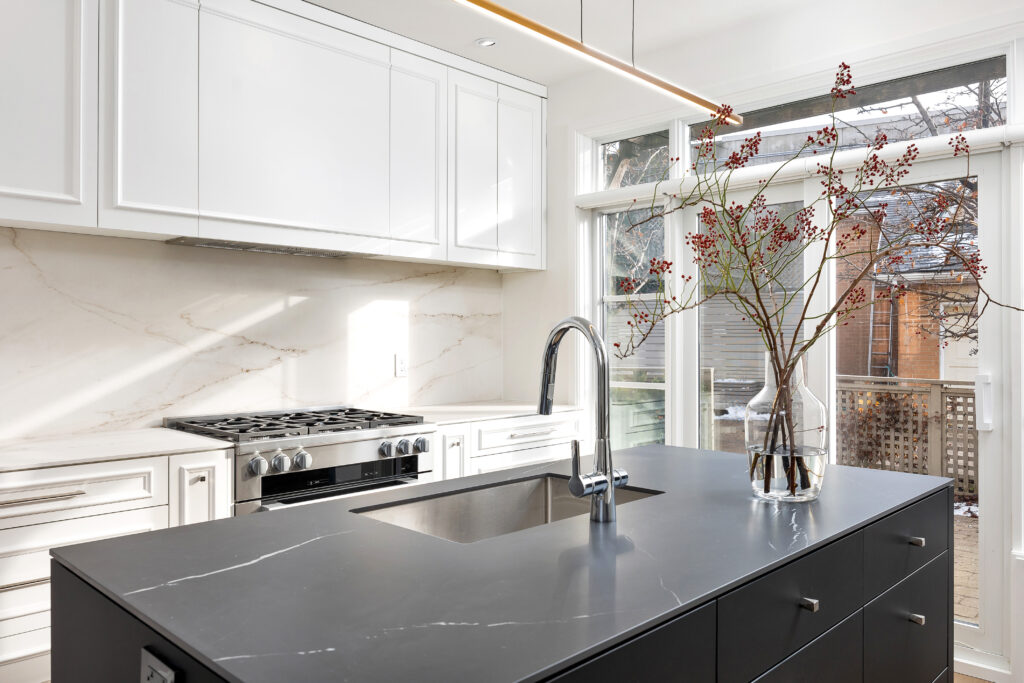 Modern kitchen with white cabinets, marble backsplash, stainless steel stove, and a black island featuring a built-in sink. A glass vase with branches decorates the countertop near large windows.