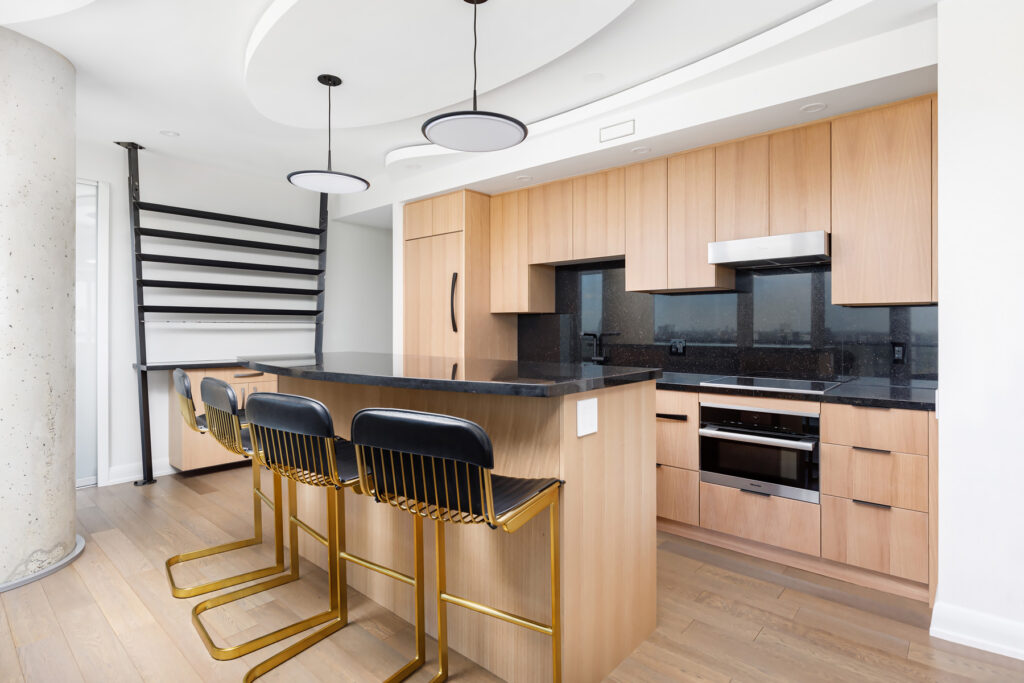 Modern kitchen with light wood cabinets, black countertops, built-in appliances, and a large island with three black and gold barstools. The space features contemporary lighting and a unique ceiling design.