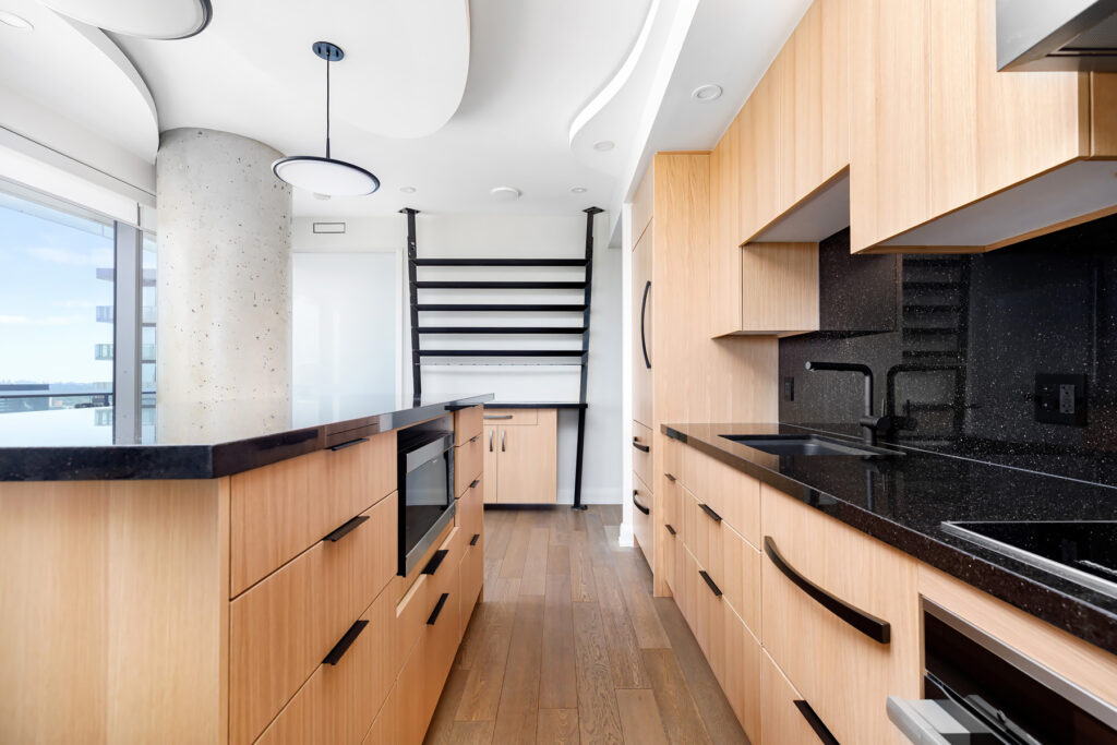 Modern kitchen with light wood cabinets, black countertops, built-in appliances, and a large island. A concrete pillar and black metal ladder accent the space, with large windows providing natural light.