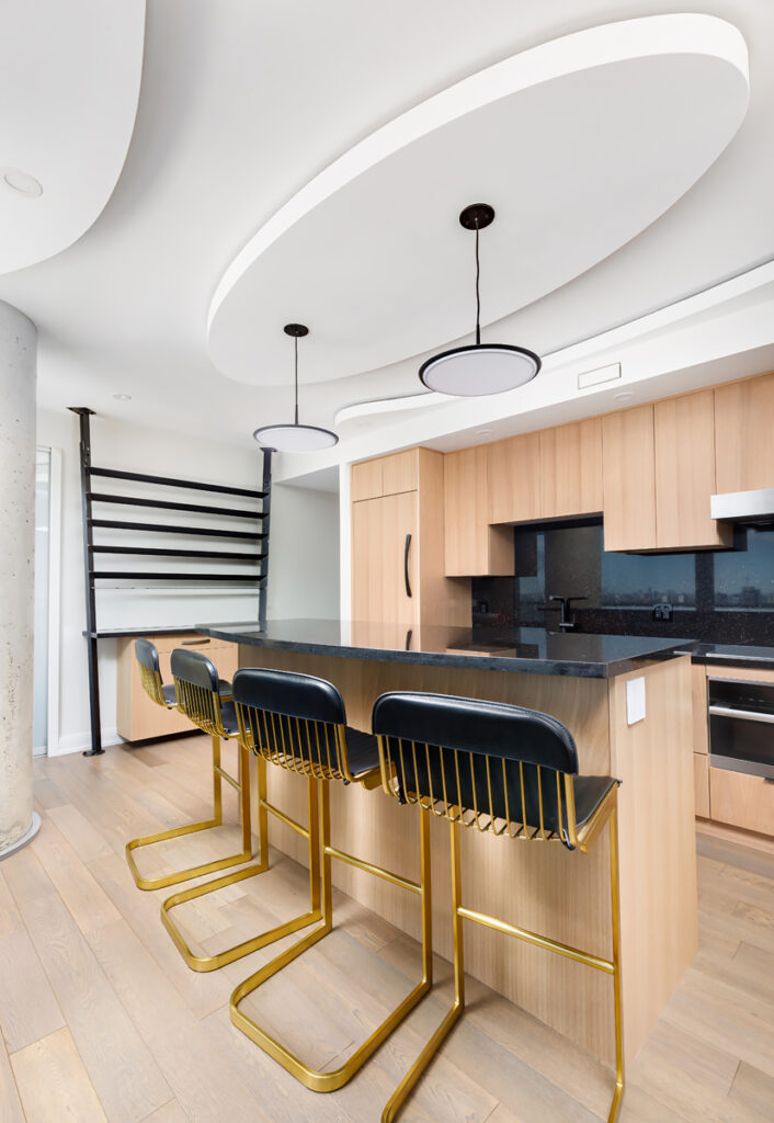 Modern kitchen with light wood cabinets, black countertops, and three black barstools with gold frames at an island. Two round pendant lights hang from the ceiling, and a large pillar stands to the left.