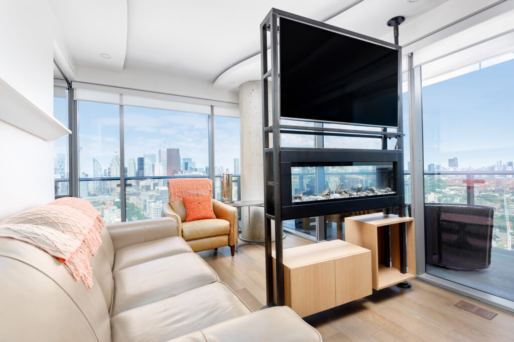 A modern living room with a beige leather sofa, an orange armchair, a large TV above an electric fireplace, and floor-to-ceiling windows showcasing a city skyline view.