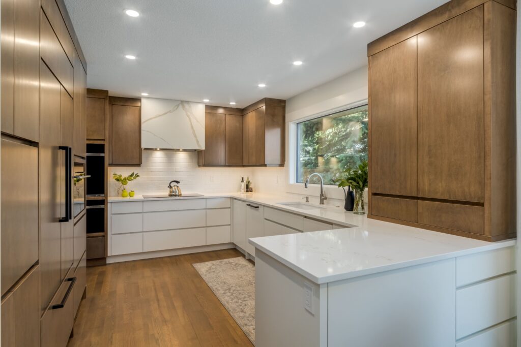 Modern kitchen with wood cabinets, white countertops, stainless steel appliances, a marble backsplash, large window, and recessed ceiling lights. There are plants and decorative items on the counters.