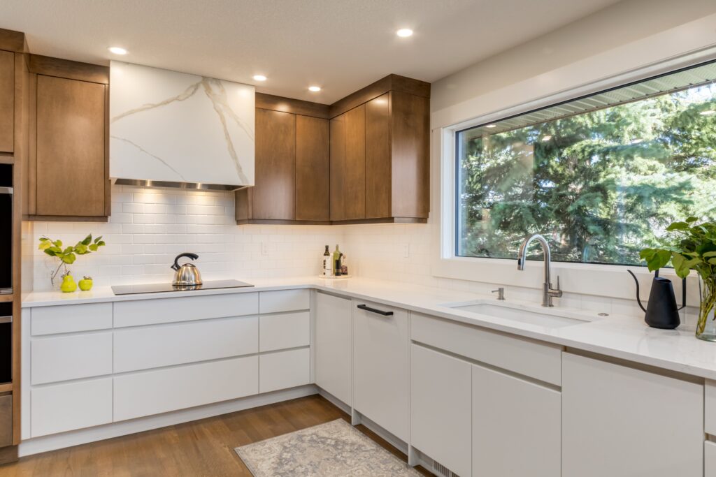 Modern kitchen with white cabinets, wood accents, a marble-patterned range hood, stainless steel appliances, a large window with outdoor view, plants, and bright lighting.