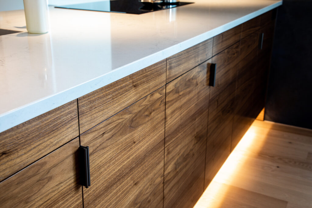 Close-up of modern kitchen cabinets with a wood grain finish, black handles, and a white countertop. Warm under-cabinet lighting highlights the texture of the wood and light-colored wooden flooring.
