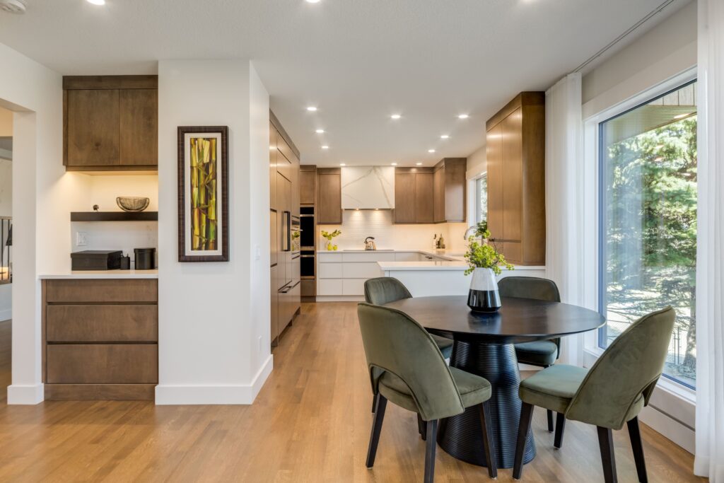 Modern kitchen and dining area with wooden cabinets, round black table, four green velvet chairs, large window with outdoor view, and decorative artwork on the wall. Bright and spacious with natural light.