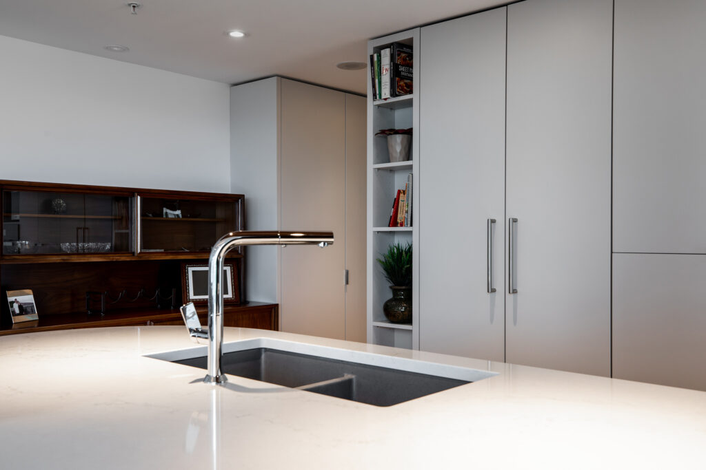 A modern kitchen with a white countertop island, built-in sink, chrome faucet, and minimalistic grey cabinets. A small open shelf with books and a plant is visible, along with a wooden cabinet in the background.