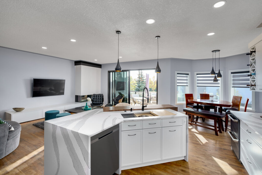 Modern open-concept kitchen and living area with white cabinets, marble island, pendant lights, large windows, dining table, and a wall-mounted TV in a bright, spacious setting with wood floors.