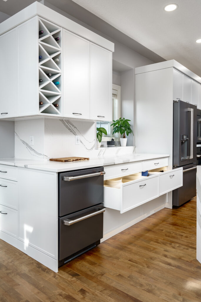 Modern white kitchen with wood flooring, black appliances, open drawers, a wine rack with bottles, marble countertops, and a potted plant near a window.