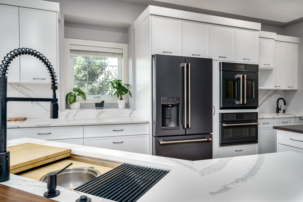 Modern kitchen with white cabinets, black appliances, a double wall oven, a French door refrigerator, a farmhouse sink with a black pull-down faucet, and green plants on the windowsill above the counter.