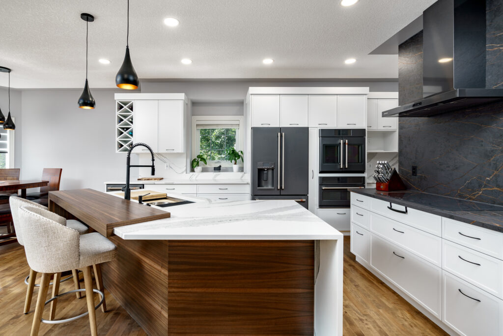 Modern kitchen with white cabinets, stainless steel appliances, a marble countertop island with wood accents, barstools, black pendant lights, and a dining area visible to the left. A window lets in natural light.