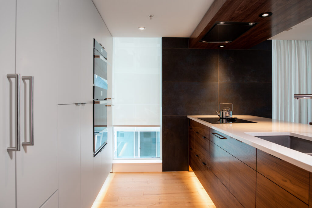 Modern kitchen with sleek white cabinets, built-in oven, dark backsplash, wooden lower cabinets, white countertops, under-cabinet lighting, and a large window letting in natural light.