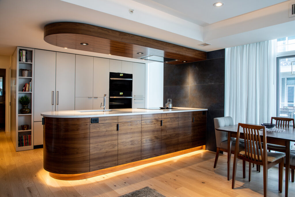 Modern kitchen with curved wood island, white countertop, built-in appliances, white cabinets, open shelving with books and a plant, dining table with chairs, large windows, and wood flooring.