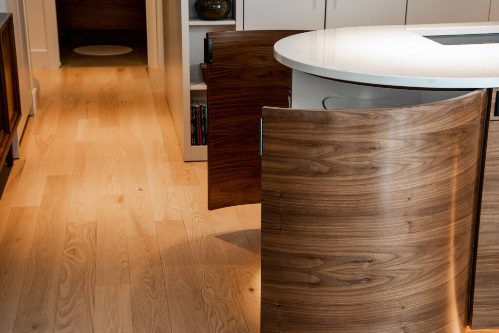 Curved wooden cabinet doors beneath a white, rounded kitchen countertop are partly open, revealing storage inside. The kitchen floor is light hardwood, and books are visible on a shelf in the background.