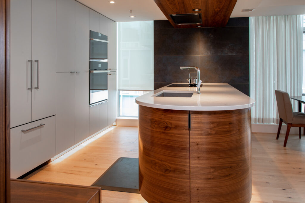 Modern kitchen with a large, curved wooden island featuring a white countertop and built-in sink. Sleek cabinets, wall ovens, wood floors, and tall windows with sheer curtains create a bright, contemporary space.
