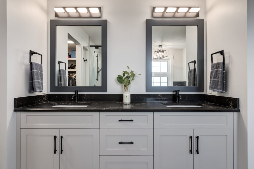 Modern bathroom vanity with double sinks, black countertops, white cabinets, two framed mirrors, and wall-mounted lights above each mirror. A vase with green leaves sits between the sinks, and hand towels hang on both sides.